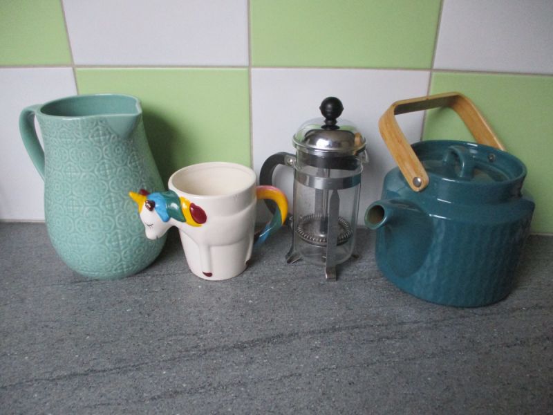 Photo de plusieurs objets sur un plan de travail de cuisine. De gauche a droite : une carafe, une tasse en forme de licorne, une cafetière et une théière
